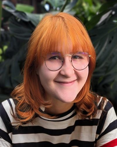 A person with copper red shoulder length hair wearing a striped shirt and glasses.