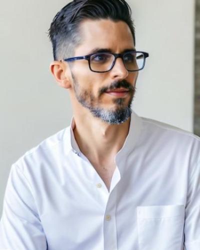man with dark hair and glasses looking to the right of the screen. He's wearing a white button down shirt with the collar unbuttoned.