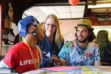 image: young adult Black male with a red shirt and wearing a blue protective helmet sitting at a table with two young adult camp counselors assisting him